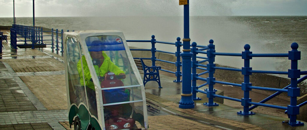 A man riding his mobility scooter, with a cover over the top, along a promenade in the rain with waves crashing against the sea wall beside him