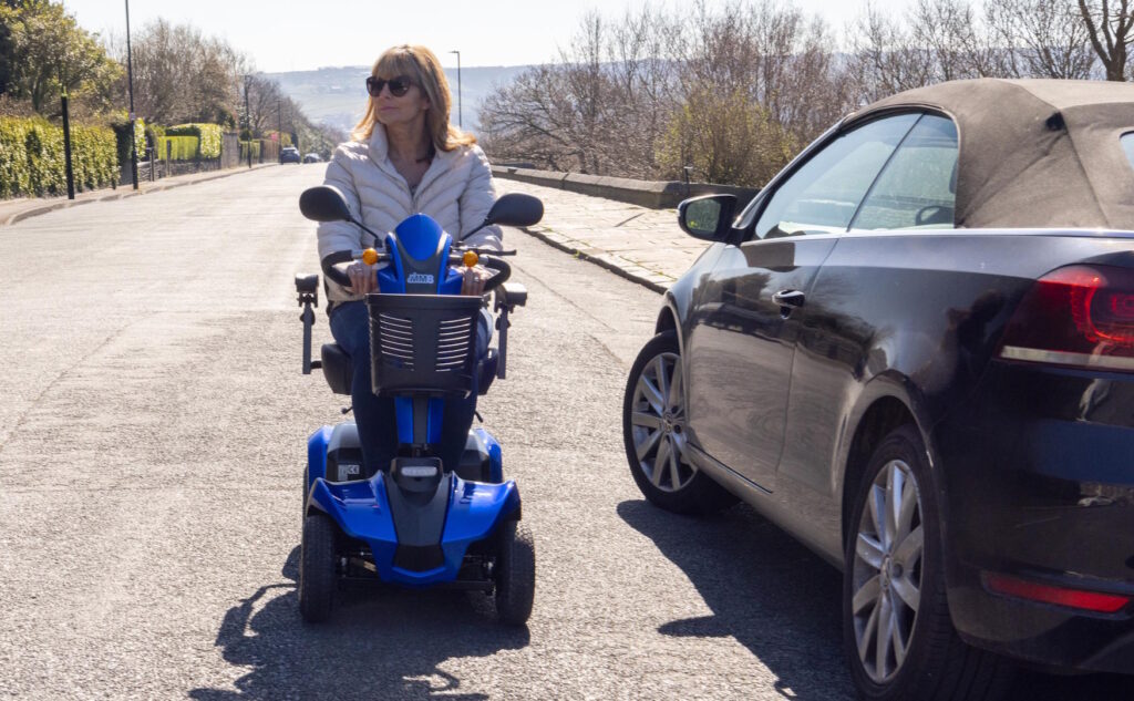 front view of a lady driving a blue MM8 mobility scooter on a road alongside a parked car